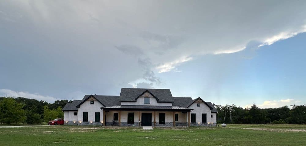 Newly constructed house with large porch, surrounded by green field under a cloudy sky.