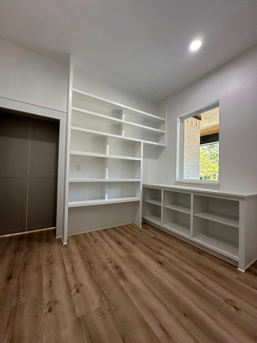 Modern empty room with built-in white shelves, large window, and hardwood flooring.