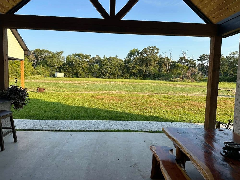 Scenic view of a green landscape from a rustic porch with wooden beams and stone flooring.