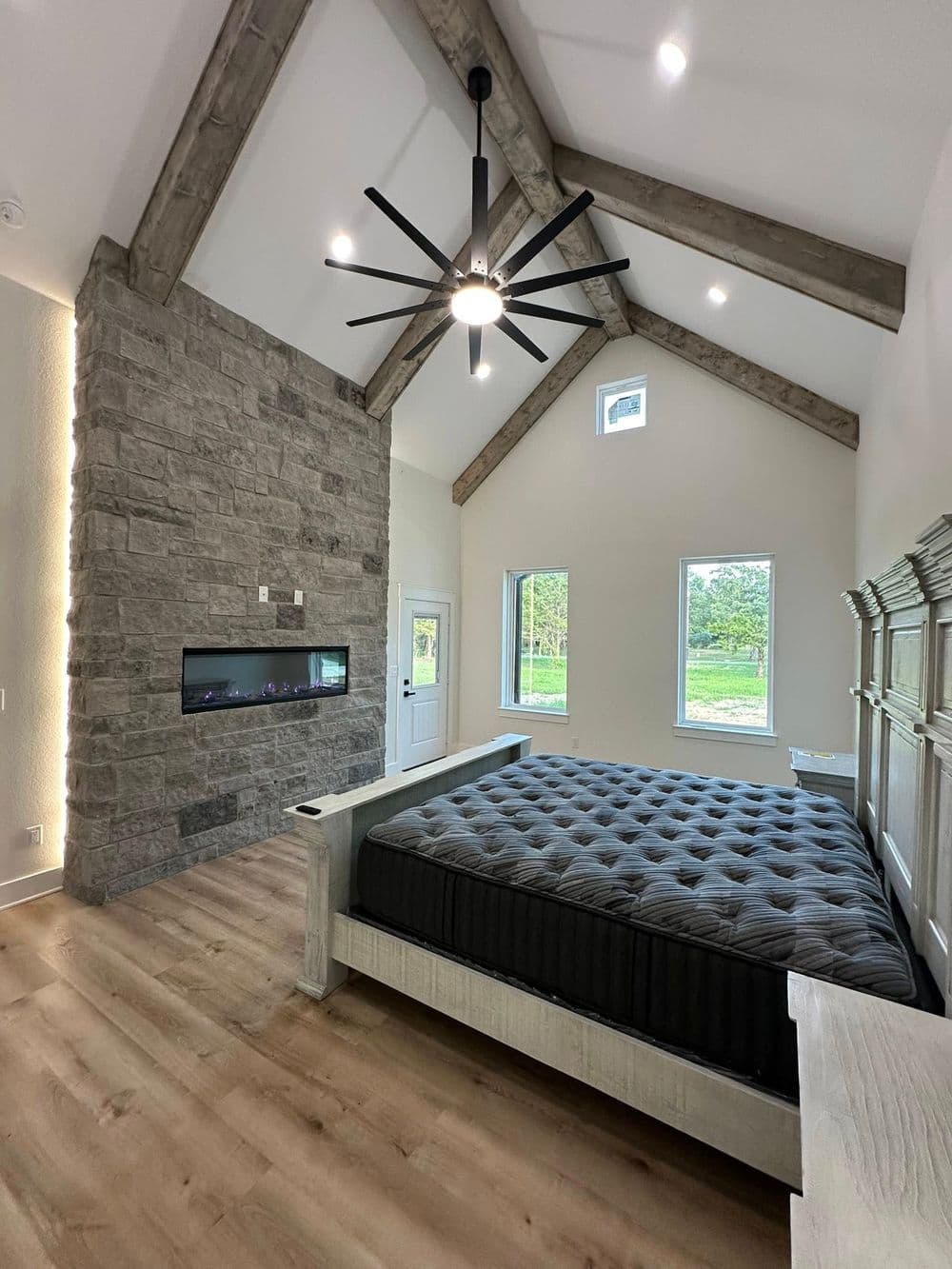 Modern bedroom with vaulted ceiling, stone accent wall, large windows, and stylish bed.