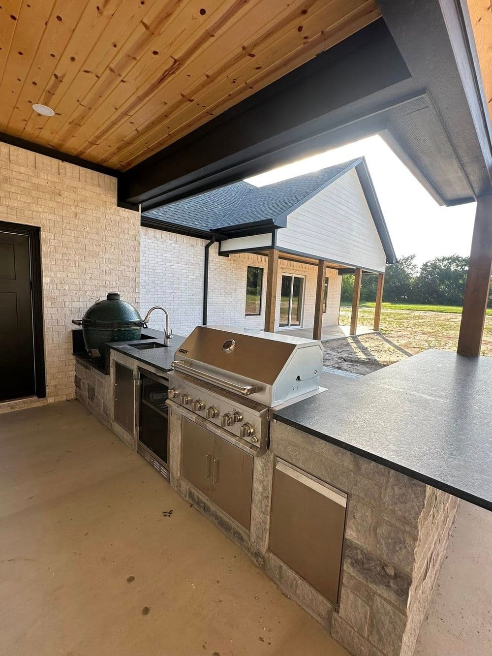 Outdoor kitchen setup with grill and stone countertops under a wooden ceiling. Modern home in background.